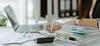 Person resting arm on ample stack of documents with laptop beside
